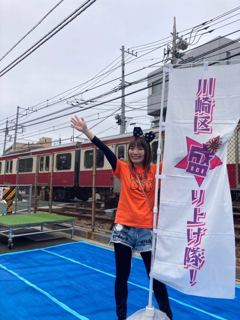 京急電鉄の車両を背景に、「川崎区盛り上げ隊！」の旗を持ってポーズをとる女性