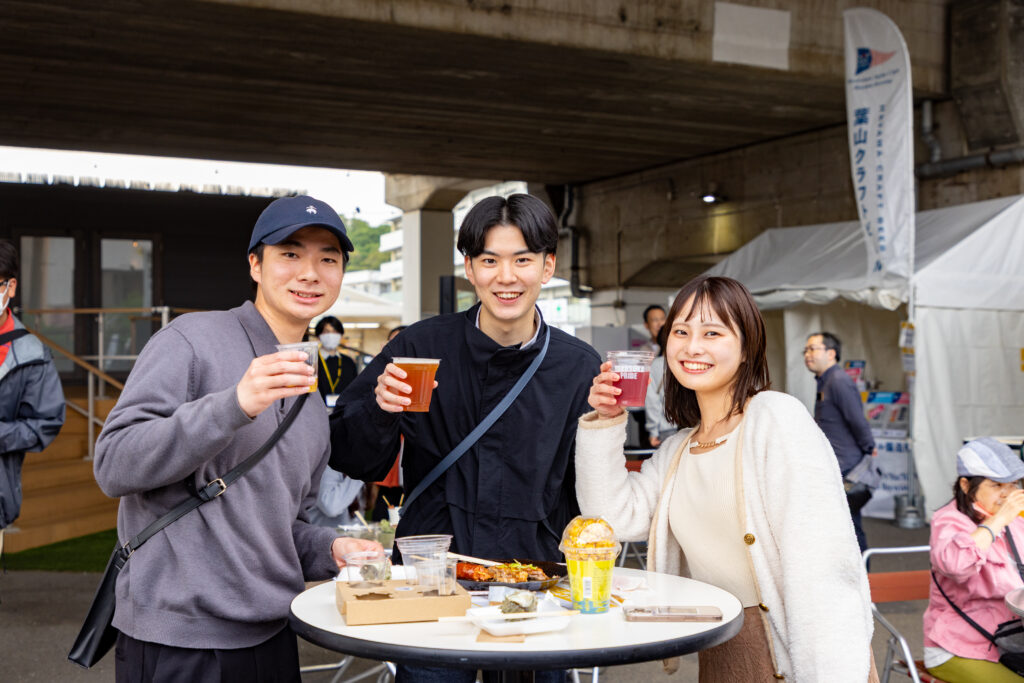 野外のテーブルでビールを持ってポーズを取る男女の若者三人